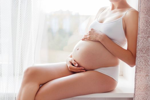 Pregnant woman in white underwear sitting on window sill. Young woman expecting a baby. Cozy happy background in sunny morning.