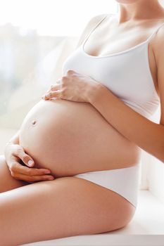 Pregnant woman in white underwear sitting on window sill. Young woman expecting a baby. Cozy happy background in sunny morning.