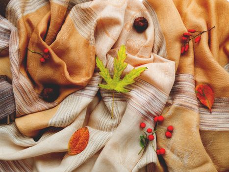 Beige scarf with fallen leaves and berries on white background. Folded warm accessory with autumn plants. Top view, flat lay.