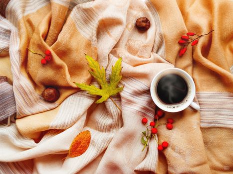 Cup of hot coffee with fallen leaves and berries on beige scarf. Folded warm accessory with autumn plants and tasty beverage. Top view, flat lay.