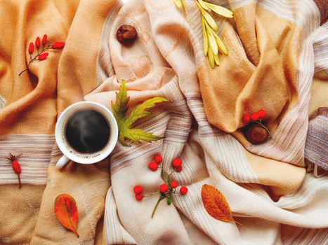Cup of hot coffee with fallen leaves and berries on beige scarf. Folded warm accessory with autumn plants and tasty beverage. Top view, flat lay.