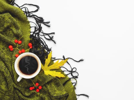 Green scarf with fallen leaves and berries on white background. White cup of hot coffee on folded warm accessory with copy space.