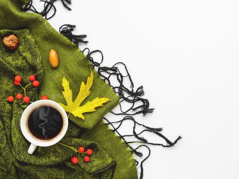 Green scarf with fallen leaves and berries on white background. White cup of hot coffee on folded warm accessory with copy space.