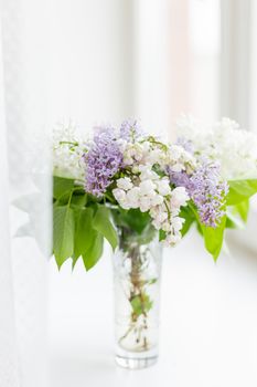 Lilac flowers in glass vase on window sill. Natural spring background with white and violet flowers.