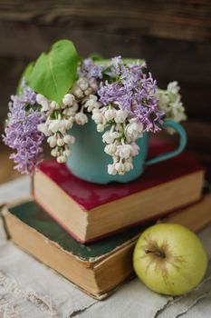 Two old books, apple and rusty mug with lilac bouquet on homespun napkin. Interesting reading, vintage background.