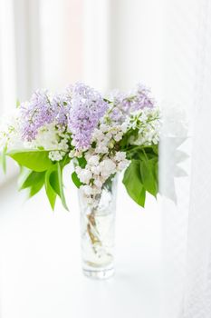 Lilac flowers in glass vase on window sill. Natural spring background with white and violet flowers.