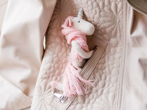 Cute fairy unicorn with a pink mane and a tail made of threads. Crocheted hand made toy in pocket of leather jacket. Trendy creature, symbol of magic and miracles.