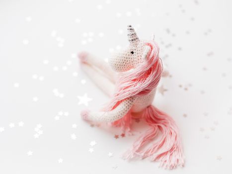 Cute fairy unicorn with a pink mane and a tail made of threads. Crocheted hand made toy on white background with silver stars confetti. Trendy creature, symbol of magic and miracles.