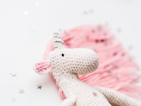Cute fairy unicorn with a pink mane and a tail made of threads. Crocheted hand made toy on white background with silver stars confetti. Trendy creature, symbol of magic and miracles.