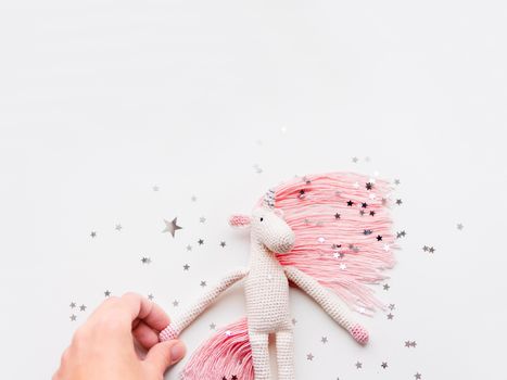 Cute fairy unicorn with a pink mane and a tail made of threads. Crocheted hand made toy on white background with silver stars confetti. Trendy creature, symbol of magic and miracles.