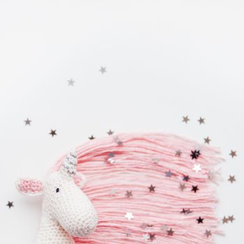 Cute fairy unicorn with a pink mane and a tail made of threads. Crocheted hand made toy on white background with silver stars confetti. Trendy creature, symbol of magic and miracles. Place for text.