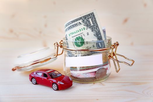 Collecting money for new car. Glass tin as moneybox with cash savings (banknotes and coins) and toy car on wooden background. White empty space for your text.