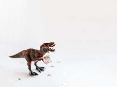 Dinosaur with present. Plastic toy with gift wrapped in silver paper. White background with star confetti. Place for text.
