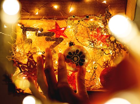 Cardboard box with Christmas and New Year decorations and light bulbs. Woman touching golden sparkling ball with flower.