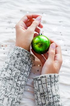 Woman holds a new year decoration - sparkling green Christmas ball. White fabric background with silver snowflakes.