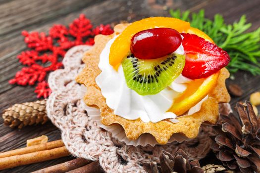 Winter holiday rustic background with fruit tart and spices - cinnamon, anise, peanuts. Christmas and New Year decorations - red snowflake, thuja and pine cones. Tasty dessert with strawberry, kiwi, orange, peach and whipped cream.