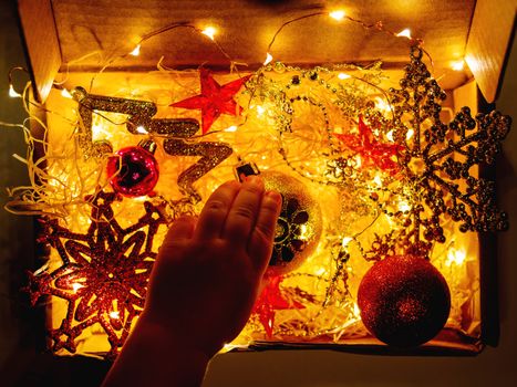 Cardboard box with Christmas and New Year decorations and light bulbs. Little baby touching golden sparkling ball with flower.