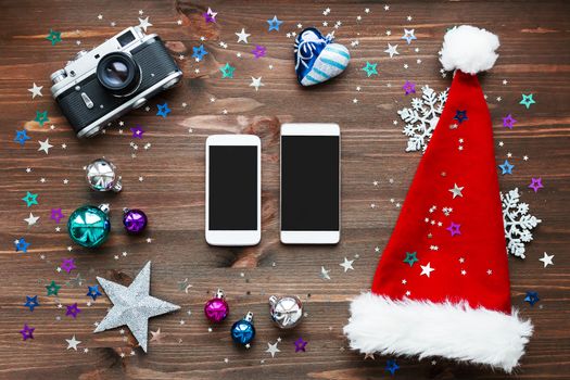 Christmas and New Year background with two smartphones, old fashioned camera, red Santa's hat, decorations - balls, stars, silver sparkling snowflakes, confetti on wooden table. Mock up, place for your text.