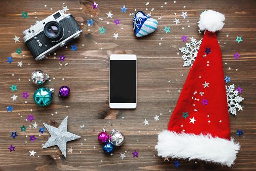 Christmas and New Year background with smartphone, old fashioned camera, red Santa's hat, decorations - balls, stars, silver sparkling snowflakes, confetti on wooden table. Mock up, place for your text.