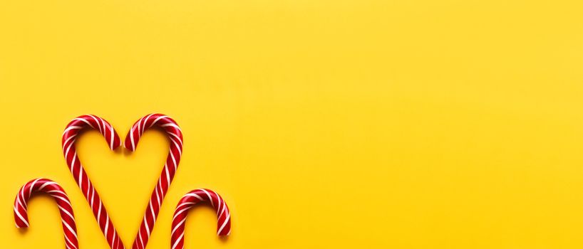 Heart made of two Christmas candy cones on yellow background. Colorful holiday sweets with bright copy space. Traditional dessert wtih red and white stripes.