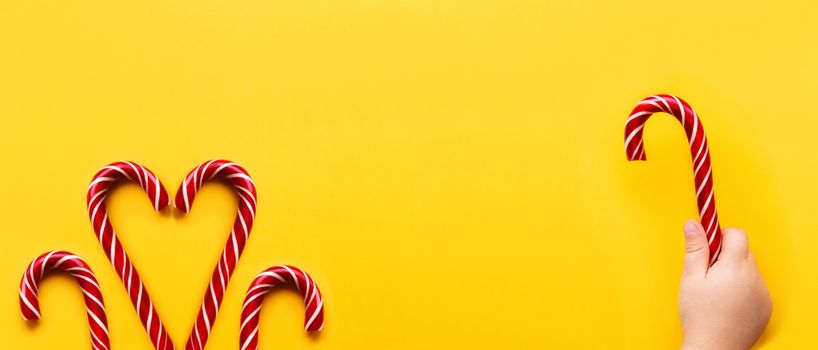 Heart made of two Christmas candy cones on yellow background. Colorful holiday sweets with bright copy space. Traditional dessert wtih red and white stripes.