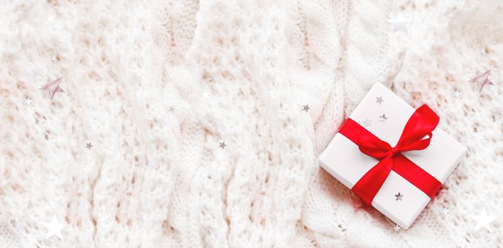 Present in white wrapping paper on white knitted backdrop. Holiday gift with red ribbon and bow. Banner with copy space.