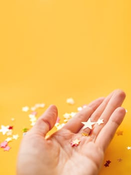 Yellow holiday background with colorful star confetti on hand. Copy space for Christmas and New Year cards.