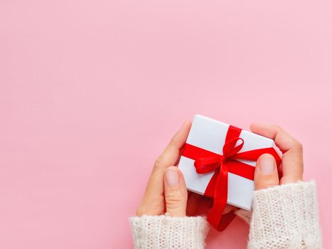 Woman holds a present in white wrapping paper on pink backdrop. Holiday gift with red ribbon and bow. Copy space.