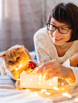 Cute ginger cat with bites light bulbs. Woman playing with her fluffy pet and box with Christmas decorations. Cozy home before New Year.