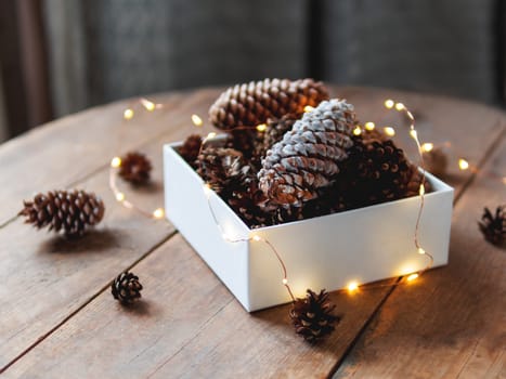 White box with pine cones and light bulbs on wooden table. Scandy style. Preparation for Christmas and New Year celebration.