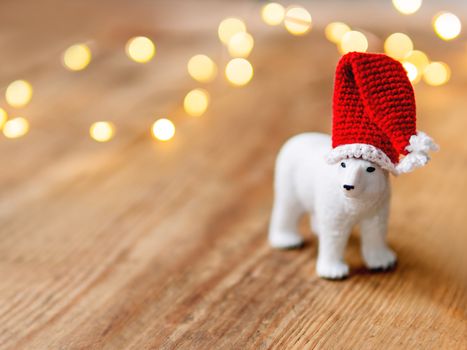 Cute polar bear in Santa's hat standing on shabby wooden table with light bulbs on background.