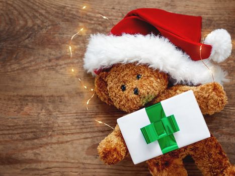 Teddy bear in Santa Claus hat. Plush toy with light bulbs on shabby wooden background. New Year present with green ribbon.
