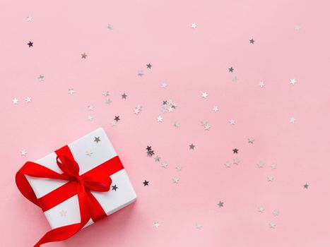 Present in white wrapping paper on pink backdrop with silver star confetti. Holiday gift with red ribbon and bow. Top view, flat lay.