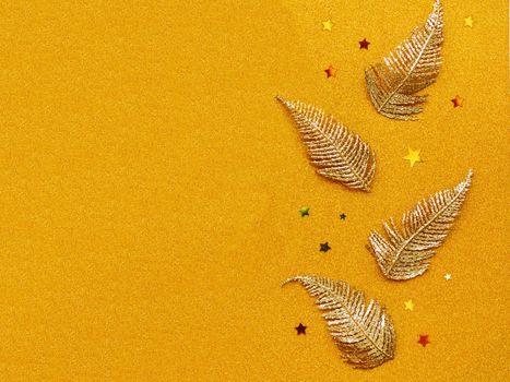 Row of golden New Year decorations on golden glitter background. Bright shiny feathers or leaves and stars confetti for Christmas tree. Top view, flat lay.