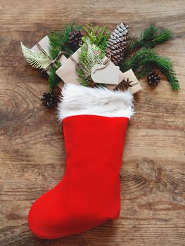Christmas presents in Santa Claus red sock. Gifts in craft paper with fir tree branches and clear tag on wooden background.