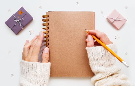 Woman is preparing to write or draw something in notepad with craft paper pages. Presents in pink and violet wrapping paper and silver stars confetti on background.