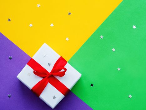 Present in white wrapping paper on colorful background. Yellow, violet and green geometrical backdrop with star confetti. Holiday gift with red ribbon and bow.