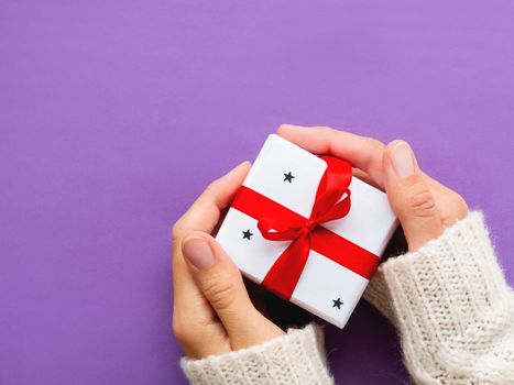 Woman holds a present in white wrapping paper on violet backdrop. Holiday gift with red ribbon and bow. Copy space.