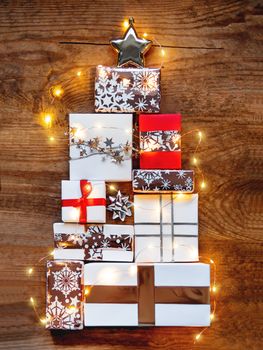 New Year presents in shape of Christmas tree on shabby wooden background. Gifts in white and golden paper with bright ribbons and light bulbs.
