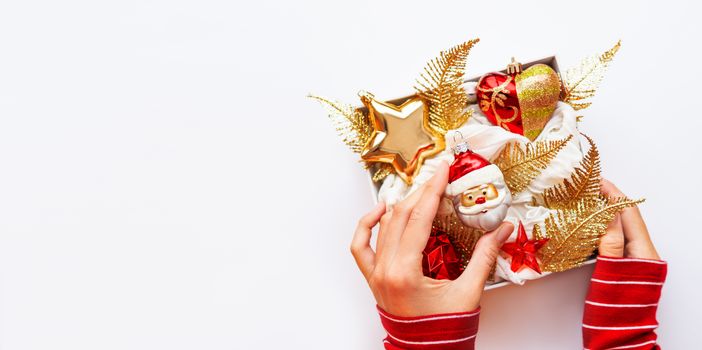 Woman in red sweater is holding white box with New Year decorations. Bright red and golden decorative toys for Christmas tree. Top view, flat lay.