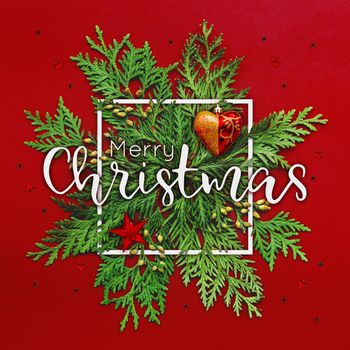 Christmas background with thuja branches and words MERRY CHRISTMAS in white square frame. Trendy Xmas greeting with stars and heart decorations on red backdrop.