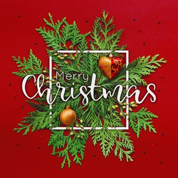 Christmas background with thuja branches and words MERRY CHRISTMAS in white square frame. Trendy Xmas greeting with stars and heart decorations on red backdrop.