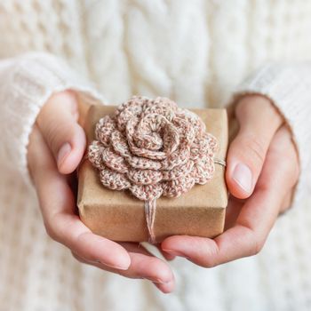 Woman in white knitted sweater holding a present wrapped in craft paper with hand made crocheted flower. DIY Cristmas or New Year gift.