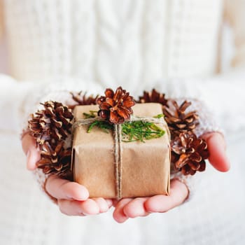 Woman in white knitted sweater holding a present packed in craft paper with pine cones. DIY Christmas or New Year, St. Valentine Day gift.