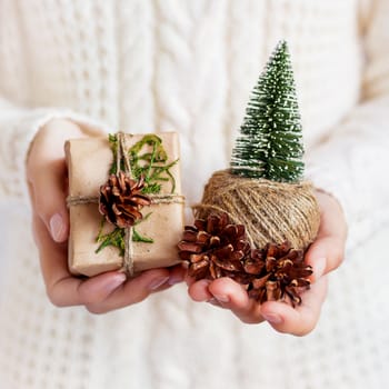 Woman in white knitted sweater holding a present packed in craft paper with pine cone. DIY Christmas or New Year, St. Valentine Day gift.