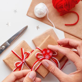 DIY presents wrapped in craft paper. Gifts tied with white and red threads and red heart. Boxes in hands on white background.