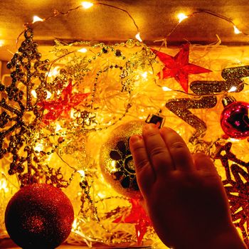 Cardboard box with Christmas and New Year decorations and light bulbs. Little baby touching golden sparkling ball with flower.