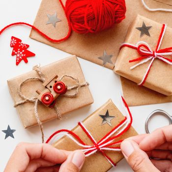Woman is packing Christmas and New Year DIY presents in craft paper. Gifts tied with threads. Boxes, red heart and Christmas tree symbols on white background.