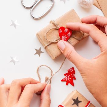 Woman is packing Christmas and New Year DIY presents in craft paper. Gifts tied with white and red threads. Boxes and red heart and Christmas tree symbols on white background.