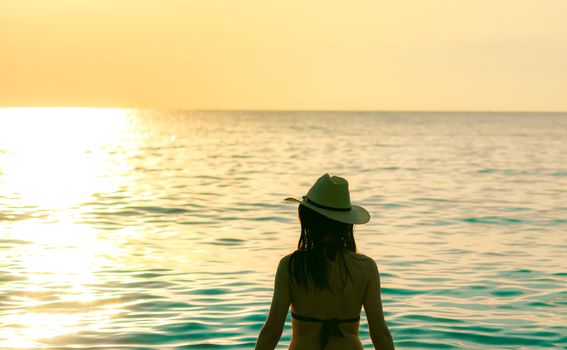 Back view of Asian woman wear bikini and straw hat sit on sand beach relax and enjoy holiday at tropical paradise beach. Woman watch beautiful sunset. Summer vacation. Travel alone. Summer vibes.